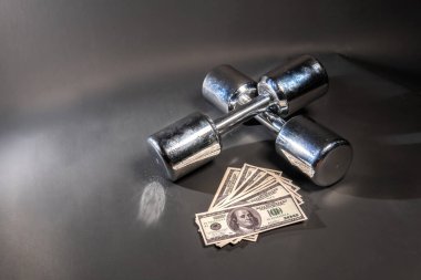 Fitness dambılları yalan söyler, para dolarları siyah arka planda gümüş renkte parıldar.