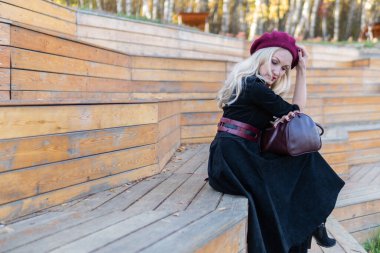 Merry bir yaz tiyatrosu bankında oturur, bordo palto ve biet ile ahşap, sonbaharda bir gölün arka planında mavi bulutlar.