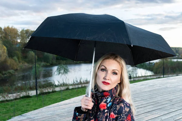 Uma menina com um guarda-chuva preto senta-se em uma escada de madeira, no fundo de uma nuvem inchada no palco. a olhar para a câmara — Fotografia de Stock