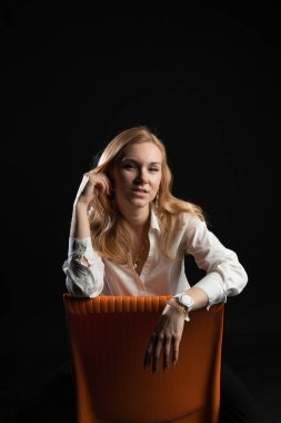 Gençler turuncu bir sandalyede oturuyorlar. Karizmatik bir görünüş, refah kavramı, stüdyodaki siyah bir arka planda, pedjak iş kadını, sarışın