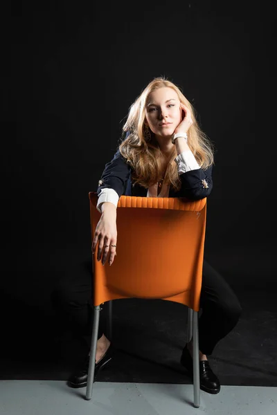 若い女の子がオレンジの椅子に座っている。カリスマ的な外観と笑顔,ウェビナー活動の概念,スタジオで黒の背景に, pedjakビジネス女性で,ブロンド — ストック写真
