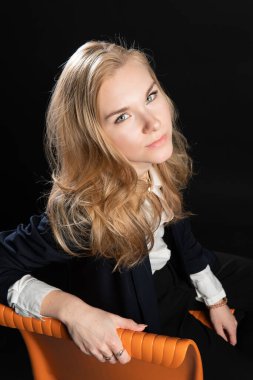 Turuncu bir sandalyede oturuyordu. Karizmatik bir görünüş, refah konsepti, stüdyodaki siyah bir arka plan, spor ceketli bir kadın, sarışın...