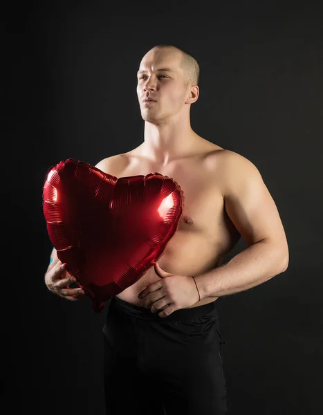 Atletik bir adam elinde kırmızı bir kalp, elinde çıplak bir balon şişirilmiş bir velentin, vücutçu romantik. Aşk adamı LGBT. Stüdyoda, siyah bir arka planda. Karizmatik ve neşeli.