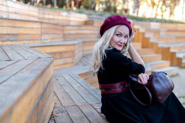 Neşeli bir kız yaz tiyatrosu bankında oturur, bordo bir palto ve biet ile ahşaptan yapılmıştır, bir yetişkin kameraya bakar, sonbaharda mavi bulutların arka planında..