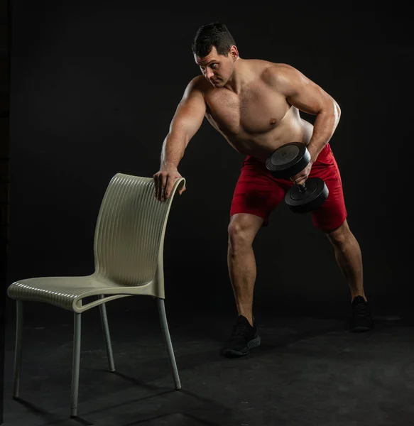 Ένας άνδρας με μια καρέκλα στο χέρι κουνάει τον δικέφαλο γυμνό κορμό του νεαρός άνδρας Καυκάσιος, μοντέρνος τύπος εμπιστοσύνης. Σοβαρή μόδα κομψότητα φόντο μαύρο bodybuilder — Φωτογραφία Αρχείου