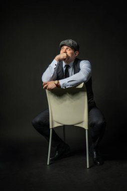 Sandalyedeki bir şapkayla genç bir erkek üç parçalı takım elbise, çekici günlük kıyafetler. Erkekler şehri, başarı gülümsemesi arka plan siyah vücut geliştirici