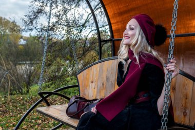 Neşeli bir bayan bahçede salıncakta oturuyor, bordo bir ceket ve biet ile dövülmüş, kameraya bakan bir yetişkin, sonbaharda bir gölün arka planında, mavi bir gökyüzü.