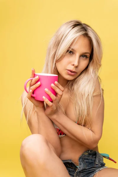 Uma menina sentada no chão com os pés cruzados mantém a cabeça em um belo fundo amarelo com uma xícara na mão amarelo ou rosa e vermelho terno de banho brincalhão sexy, figura cabelo olhando pessoa, quente — Fotografia de Stock