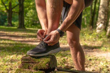 Sporcu, bacaklarının bağcıklarını spor ayakkabılarına bağlıyor. Parkta koşan sporcu, açık havada, ormanın etrafında koşuyor. Meşe ağaçları, yeşil çimenler genç sporcu, spor koşucusu.