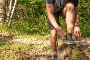 Sporcu, bacaklarının bağcıklarını spor ayakkabılarına bağlıyor. Parkta koşan sporcu, açık havada, ormanın etrafında koşuyor. Meşe ağaçları, yeşil çimenler genç sporcu doğasına dayanan spor sporu, egzersiz.