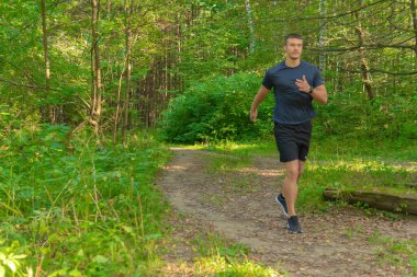 Parkta koşan bir sporcu, ormanın etrafında, meşe ağaçları, yeşil çimenler genç sporcu egzersizleri, fitness koşucusu hareketleri, ağaçlar. Yaz tatili koşusu, ayaklar esneme