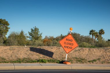 Parlak turuncu elmas şekilli Yol Kapalı Yol tabelası. Tepesinde bir ışık var. Yaya beton kaldırımın kenarında ve küçük toprak bir tepenin kenarında.