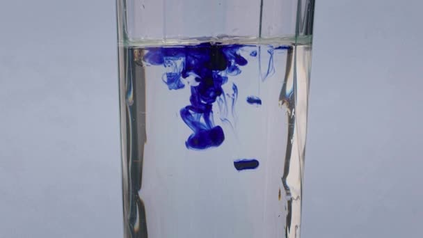 Baum kleine Tropfen hellblauer Tinten tropfen in reines Wasserglas, das auf weißem Hintergrund steht. Langsames Mischen von Flüssigkeiten. — Stockvideo