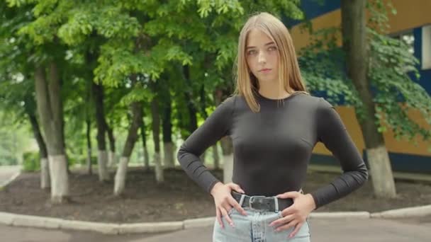 Молодая красивая девушка модели внешний вид стоит на фоне деревьев и здания, смотрит на камеру, Prores422HQ — стоковое видео