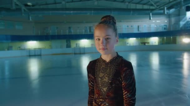 Una niña, patinadora artística, se para en la pista, mira a la cámara, la niña aprende a patinar. Prores422 — Vídeo de stock