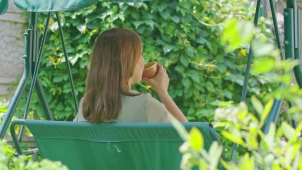 Молодая девушка летом пьет горячий чай, сидя на качелях, в своем дворе, чай помогает в жару больше всего, Prores422 — стоковое видео
