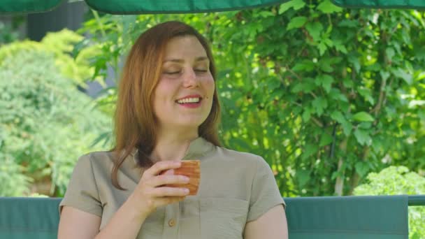 Uma jovem no verão bebe chá quente, sentado em um balanço, em seu quintal, chá ajuda no calor melhor de tudo, Prores422 — Vídeo de Stock