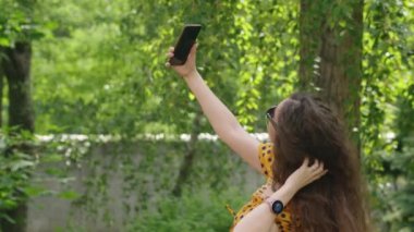 Reklam çekimi, genç bir kız gülümsüyor, telefondan yaprakları süzüp bakıyor, yazın, güzel bir elbiseyle. Dualar