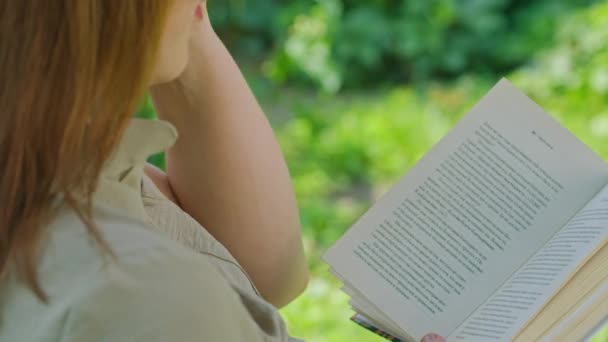 Διαφημιστικό υλικό, ένα νεαρό κορίτσι το καλοκαίρι στον κήπο διαβάζει ένα βιβλίο, κάθεται σε μια κούνια. — Αρχείο Βίντεο