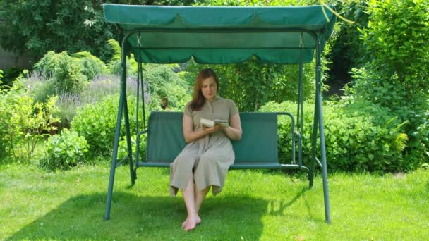 Reklámfilm, egy fiatal lány nyáron a kertben olvas egy könyvet, ül a hintán. — Stock videók