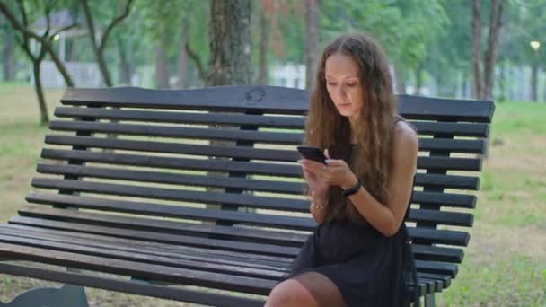 在商业拍摄中，公园里一个穿着黑色连衣裙的小女孩坐在长椅上，一边打电话一边离开. — 图库视频影像