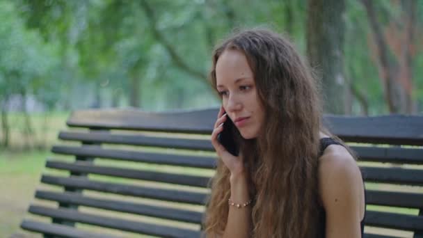Tiro comercial, uma jovem no parque se senta em um banco em um vestido preto e fala ao telefone. — Vídeo de Stock