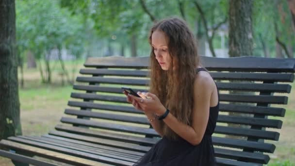 Tiro comercial, uma jovem no parque se senta em um banco em um vestido preto e folheia através do telefone. — Vídeo de Stock