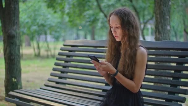 Tiro comercial, uma jovem no parque se senta em um banco em um vestido preto e folheia através do telefone. — Vídeo de Stock