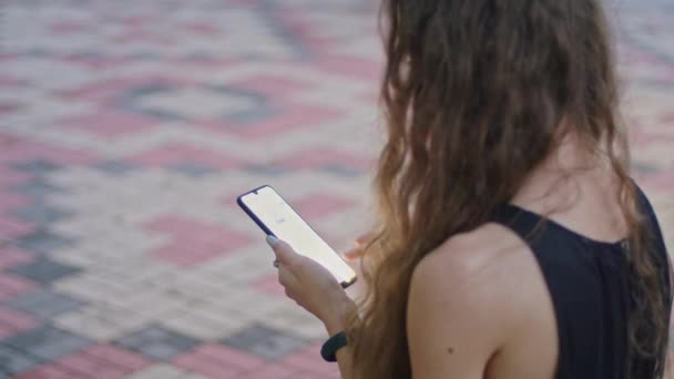 Tiroteo comercial, una joven en el parque se sienta en un banco con un vestido negro y hojas a través del teléfono. — Vídeo de stock