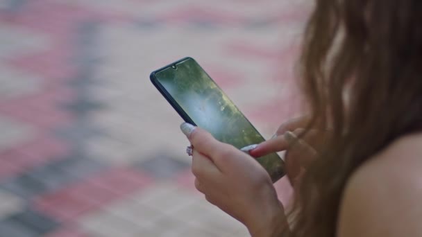 Werbedreh, ein junges Mädchen im Park sitzt in einem schwarzen Kleid auf einer Bank und blättert durchs Telefon. — Stockvideo