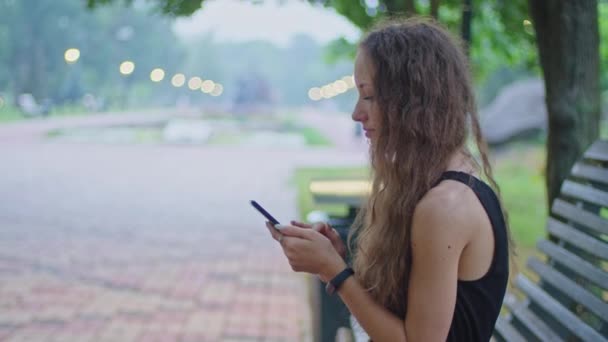 Tiro comercial, uma jovem no parque se senta em um banco em um vestido preto e folheia através do telefone. — Vídeo de Stock