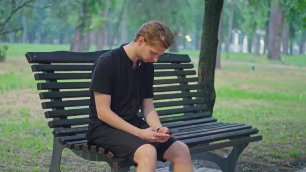 Tournage de publicité, un jeune homme dans le parc est assis sur un banc en vêtements noirs et joue au téléphone. — Video