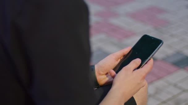 Ein junger Mann im Park sitzt in schwarzer Kleidung auf einer Bank und spielt am Telefon. — Stockvideo