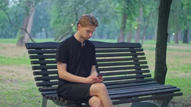 Tirando publicidad, un joven en el parque se sienta en un banco con ropa negra y juega en el teléfono. — Vídeos de Stock