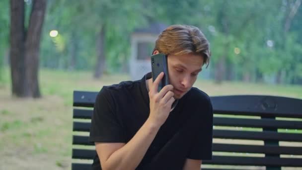 Natáčení reklamy, mladý muž v parku sedí na lavičce v černém oblečení a mluví do telefonu. — Stock video