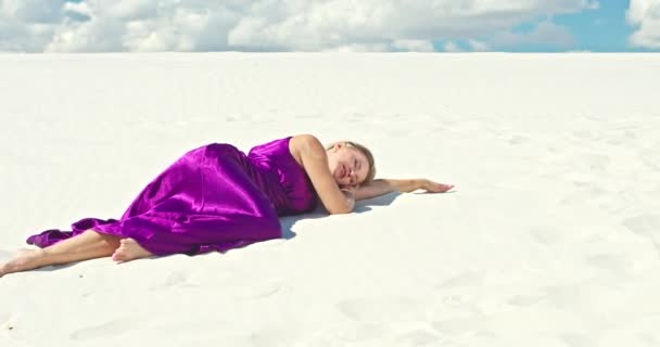砂の砂丘に寝そべっている女性の映画的なスローモーション。背景に雲と砂の砂漠の丘陵地の表面に風に揺れるドレスで裸足の女性旅行者。4K景観自然 — ストック動画