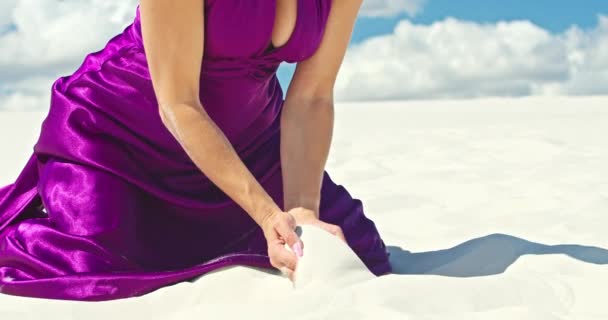 Cinematic slow motion of a woman who drew sand from her hands while sitting in the desert. Barefoot woman traveler in dress swaying in the wind on the hilly surface of the sandy desert with a cloud in — Stock Video