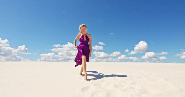 Kum tepeciğinde yürüyen bir kadının sinematik yavaş çekimi. Çıplak ayaklı kadın gezgin rüzgarda sallanan bir elbiseyle kumlu çölün dalgalı yüzeyinde arka planda bir bulutla. 4K Manzarası — Stok video
