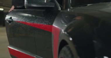 Siyah takım elbiseli, beyaz gömlekli, kırmızı kravatlı güzel bir sarışın yeraltı otoparkında siyah arabasından iniyor..