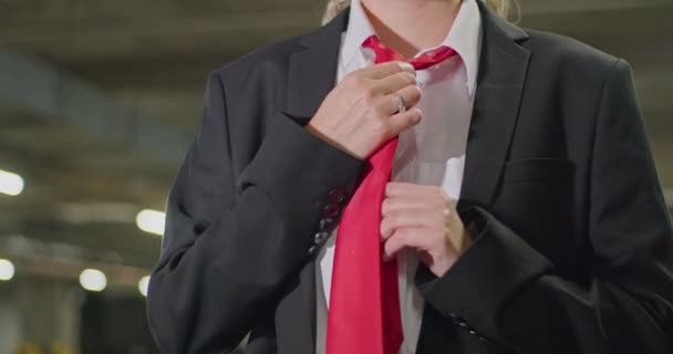 Una hermosa rubia con un traje negro, una camisa blanca endereza una corbata roja, mira a su alrededor en un estacionamiento subterráneo, una toma cinematográfica, cámara lenta. — Vídeo de stock