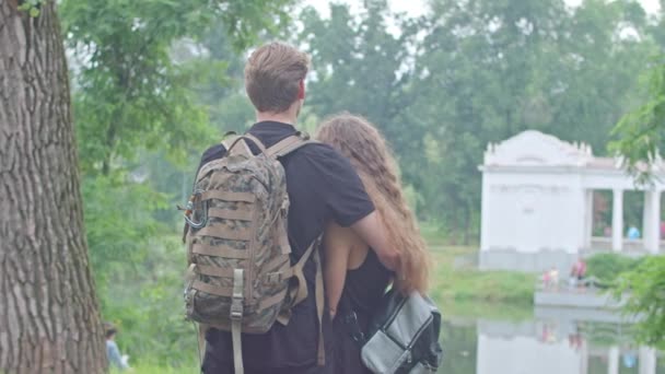 Mladý pár, muž s maskovacím batohem a dívka stojí na nábřeží, nábřeží v parku, objímají se, projevují city k sobě navzájem. — Stock video