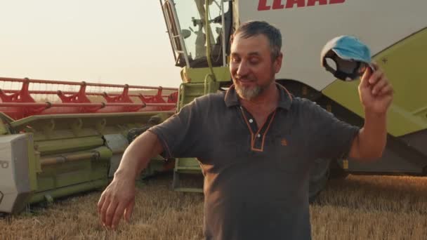 Boer staat tegen de achtergrond van een tarweveld en een maaidorser, oogsten, zonsondergang, agrarische industrie. — Stockvideo