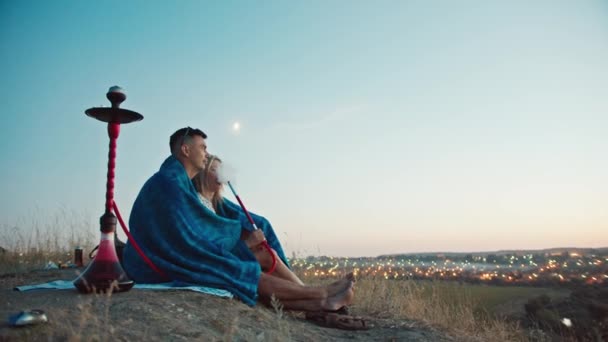 Un tipo y una chica en las montañas miran el atardecer, envueltos en una manta, fuman una cachimba, se sientan en un abrazo. Mira las luces nocturnas de la ciudad. descanso dentro de la ciudad. — Vídeos de Stock