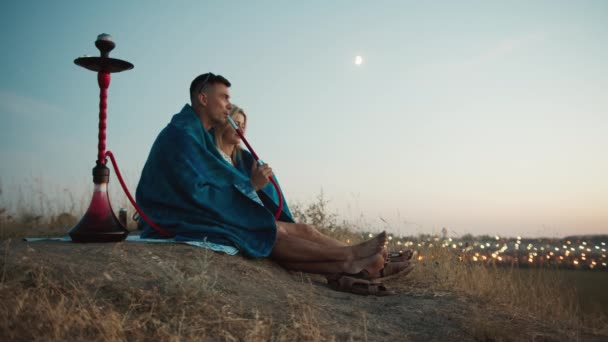 Un chico y una chica disfrutan de la puesta de sol y una vista de la ciudad, fuman una cachimba, se sientan en un abrazo, disfrutando de la vista de la ciudad. — Vídeo de stock