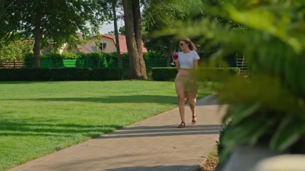 Uma bela jovem mulher em uma camiseta branca desfruta de um maravilhoso coquetel no verão quente, caminha ao longo de um beco em um lugar fresco e bonito, — Vídeo de Stock