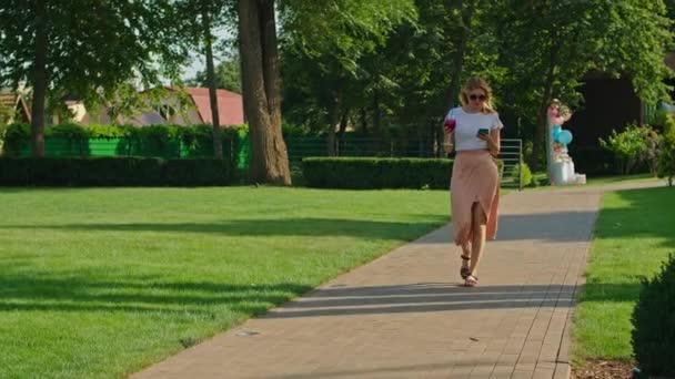 Μια όμορφη νεαρή γυναίκα με λευκό μπλουζάκι απολαμβάνει ένα υπέροχο κοκτέιλ το ζεστό καλοκαίρι, περπατά σε ένα δρομάκι σε ένα δροσερό και όμορφο μέρος, — Αρχείο Βίντεο