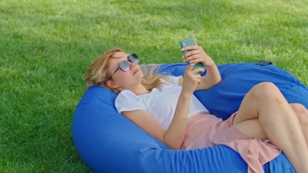 A young woman enjoys talking on the phone, texting with someone, lying on a large blue ottoman on the grass in a wonderful place, wearing a white t-shirt. — Stock Video