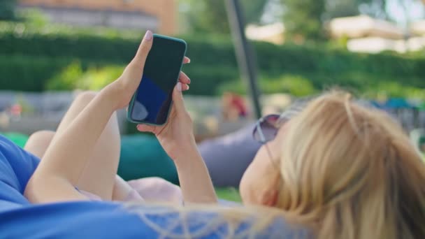 A una giovane donna piace parlare al telefono, messaggiare con qualcuno, sdraiata su un grande pouf blu sull'erba in un posto meraviglioso, indossando una t-shirt bianca. — Video Stock