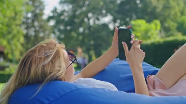 Uma jovem mulher gosta de falar ao telefone, mensagens de texto com alguém, deitado em um grande otomano azul na grama em um lugar maravilhoso, vestindo uma camiseta branca. — Vídeo de Stock
