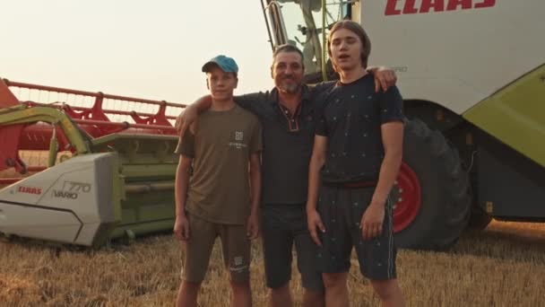 Krivoy Rog, Oekraïne - 07.18.2021 boer en zijn zonen staan tegen de achtergrond van een maaidorser in het veld, agrarische industrie, oogst, zomer. zonsondergang. — Stockvideo